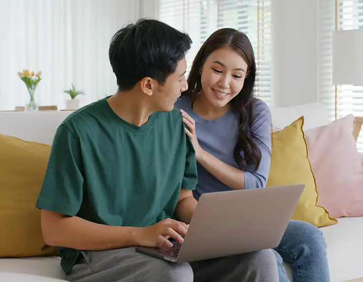 Asia people young family man woman smile happy fun playful laugh sit at home sofa couch pay for hotel booking, search choose, buy ticket on laptop social media app travel abroad trip getaway plan tour