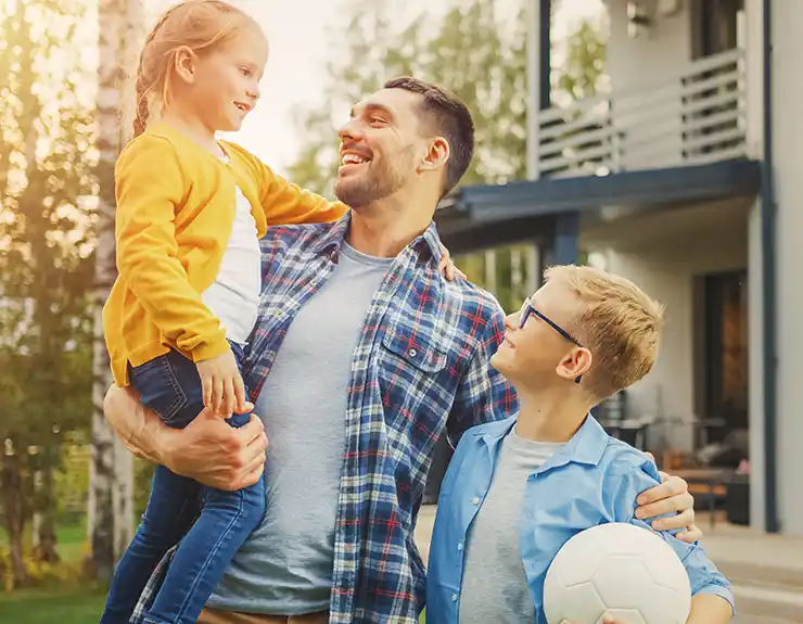 Portrait of a Happy Family of Three: Father, Daughter, Son. They Are Posing In Front of Camera on a Lawn Next to Their Country House. Dad is Holding the Girl in His Arms. Boy is Holding a Football.