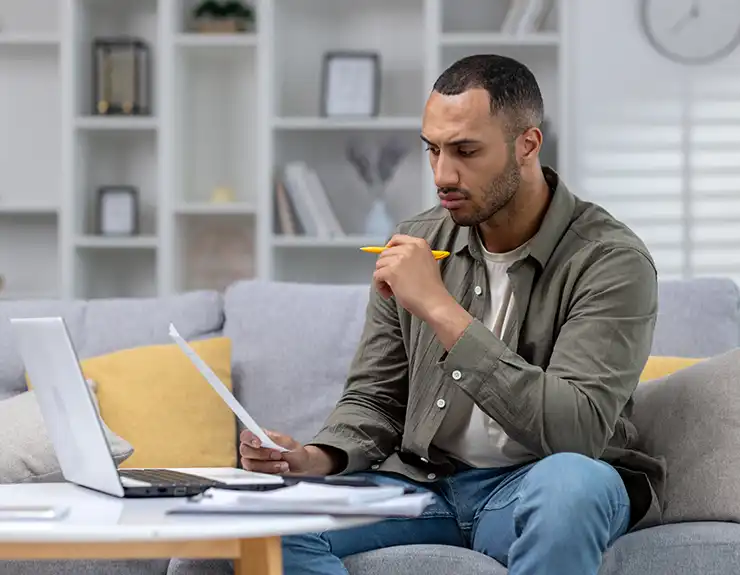 Worried and serious young African American man working at home with documents and bills. He sits huddled on the sofa in front of the laptop and holds papers in his hands