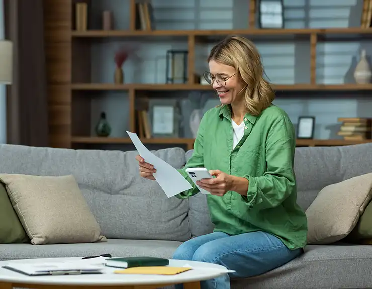 An elderly woman received good news, financial deal, loan repayment, house sale, pension, subsidy. He is sitting on the sofa at home, holding the phone and documents in his hands. Checks accounts.