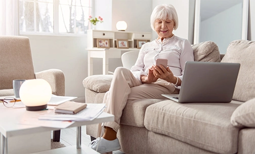 Older woman sitting on coach viewing her mobile phone and laptop.