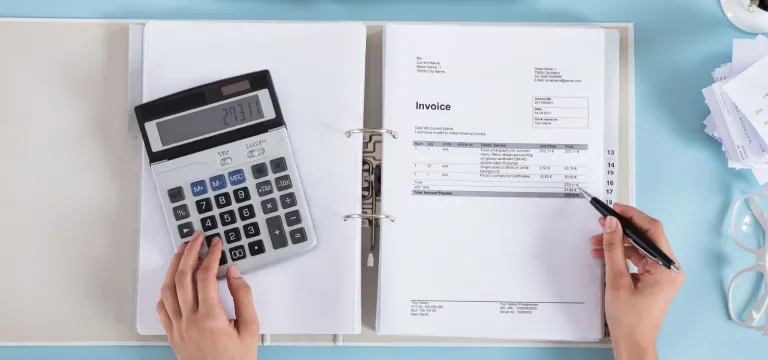 Two hands in frame, holding pen in one hand and working on calculator, invoice sheet, laptop and other stationary materials on the table.