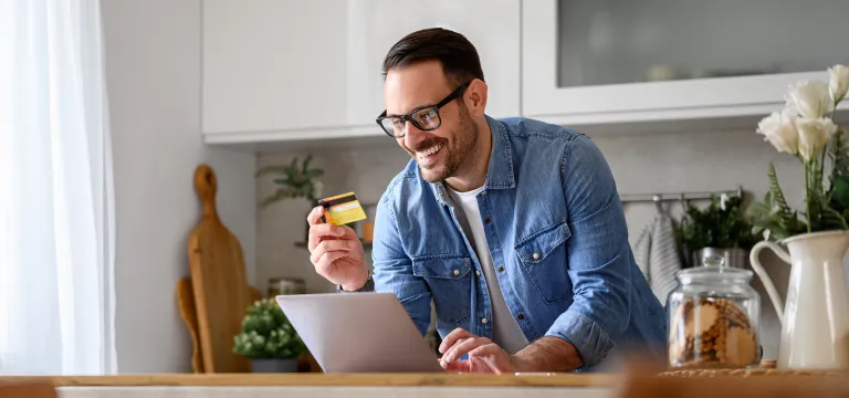 Man standing near the pantry counter top, looking at laptop screen and credit card in hand