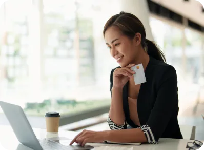  smiling businesswoman at a desk holds a card while typing on a laptop computer to shop online.