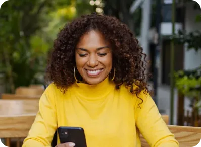 A smiling woman with curly hair sits at an outdoor cafe table looking at the PenFed mobile banking app on her phone.