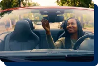 A woman sits in a convertible car with an open roof. She looks into the rearview mirror and smiles.