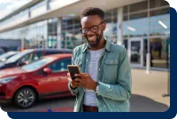 Car buyer smiling while looking at his mobile device in front of an auto dealership