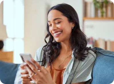 A happy woman relaxes on a blue couch at home while scrolling through her PenFed financial accounts on a smartphone.