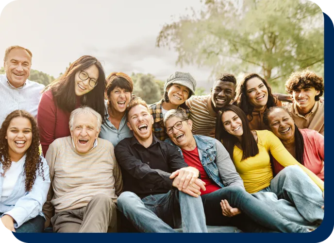 A large, diverse group of smiling people of various ages and backgrounds sitting closely together outdoors.