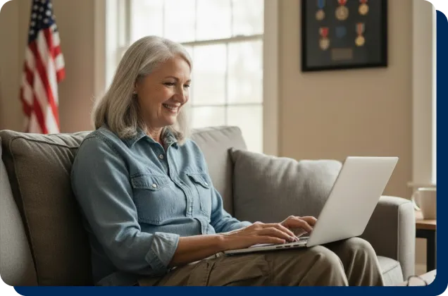 Smiling senior woman with gray hair using a laptop on a couch at home, with an American flag and military medals visible in the background.