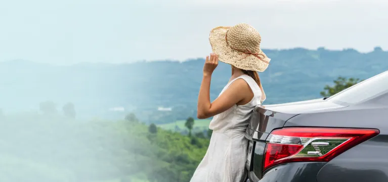 A woman in a straw hat and white dress leans against a silver car, looking out at a scenic mountain landscape.