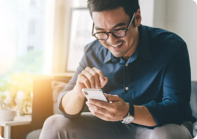 A smiling man with glasses looks at his smartphone while sitting down, likely interacting with a mobile app.