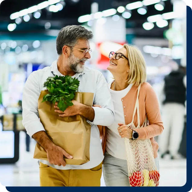 Smiling mature couple walking together while holding grocery bags in a brightly lit market. 