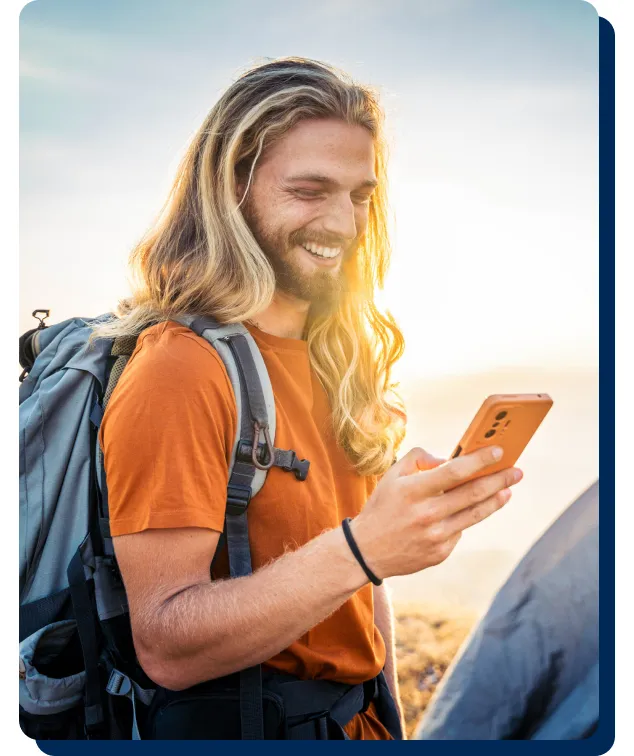 Smiling hiker with backpack looks at smartphone outdoors, illustrating PenFed personal loans for travel and adventure.