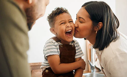 Happy boy getting a kiss by caring mother, bonding and laughing during family time at home. Young parents sharing a sweet moment of parenthood with their playful child, relaxing and carefree together